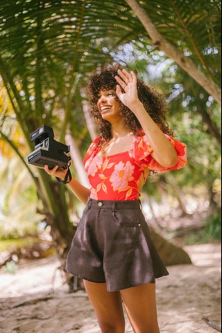 Smiling woman in floral top and black shorts holding instant camera under palm trees.